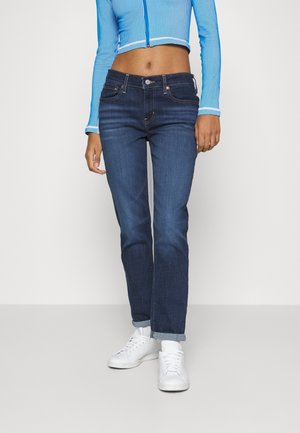 Mujer con jeans azul oscuro de pierna recta, zapatillas blancas y una camiseta azul cropped con cremallera y mangas largas, de pie sobre un fondo blanco.