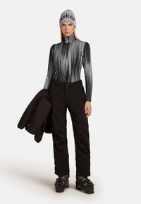 Schwarze Skihosen kombiniert mit einem grauen langärmligen Thermoshirt mit vertikal gestreiftem Muster und einer grauen Mütze mit Text. Eine schwarze Jacke wird gehalten.
