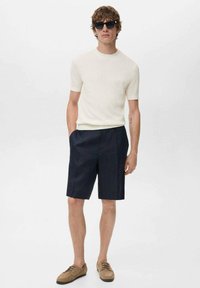 Suéter blanco de manga corta acanalado, pantalones cortos oscuros de sastre en azul marino y zapatos marrones de cordones. Modelo con gafas de sol negras. Diseño simple y moderno.