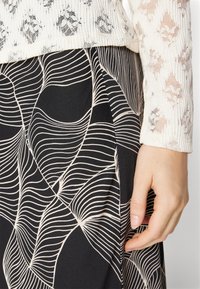 Gonna nera con motivo a onde bianche, abbinata a un top chiaro e strutturato che presenta un design simile a pizzo. Vista ravvicinata di una mano appoggiata sulla gonna.