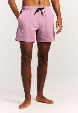 Homme portant un short de bain violet clair avec un cordon noir, debout pieds nus devant un fond blanc uni.