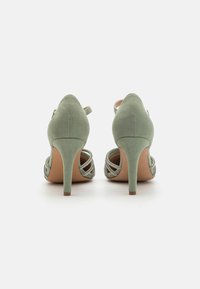 Escarpins en suède vert avec un dos ouvert et des brides croisées avec des accents métalliques. Talon fin et surface texturée.