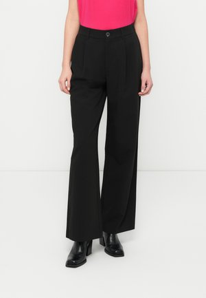 Person som har på seg høytlivede sorte vide bukser, svarte støvler med hæl og en innstikket knallrosa ermeløs topp, står foran en hvit bakgrunn.