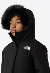 Μαύρο μονωμένο μπουφάν με κουκούλα επενδεδυμένη με γούνα, με φερμουάρ στο μπροστινό μέρος και κεντημένο λογότυπο στο αριστερό στήθος. Ομαλή υφή υφάσματος.