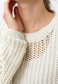 Pull en tricot léger couleur blanc cassé, présentant un design aéré et loose, un col et des poignets côtelés, avec une finition texturée.