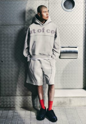 Fiatal férfi borotvált fejjel, szürke kapucnis pulóvert és rövidnadrágot visel, piros zokniban és fekete cipőben, kezét a zsebében tartva fémfal előtt áll.