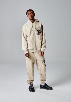 Sudadera beige con diseño gráfico, acompañada de pantalones ajustados a juego. Presenta un bolsillo tipo canguro y puños acanalados. Combinada con zapatillas negras.
