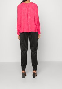 Rosa Bluse mit langen Ärmeln und gerafftem Ausschnitt, kombiniert mit schwarzen, taillierten Hosen und Sandalen mit Blockabsatz und Riemchen.