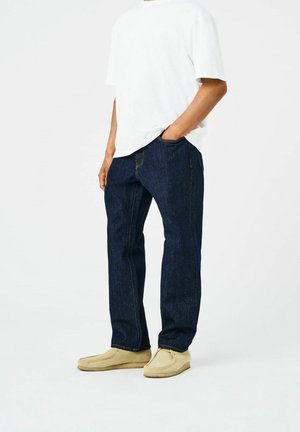 Dark blue denim jeans with straight legs, featuring orange stitching and side pockets. Paired with a plain white T-shirt and beige shoes.