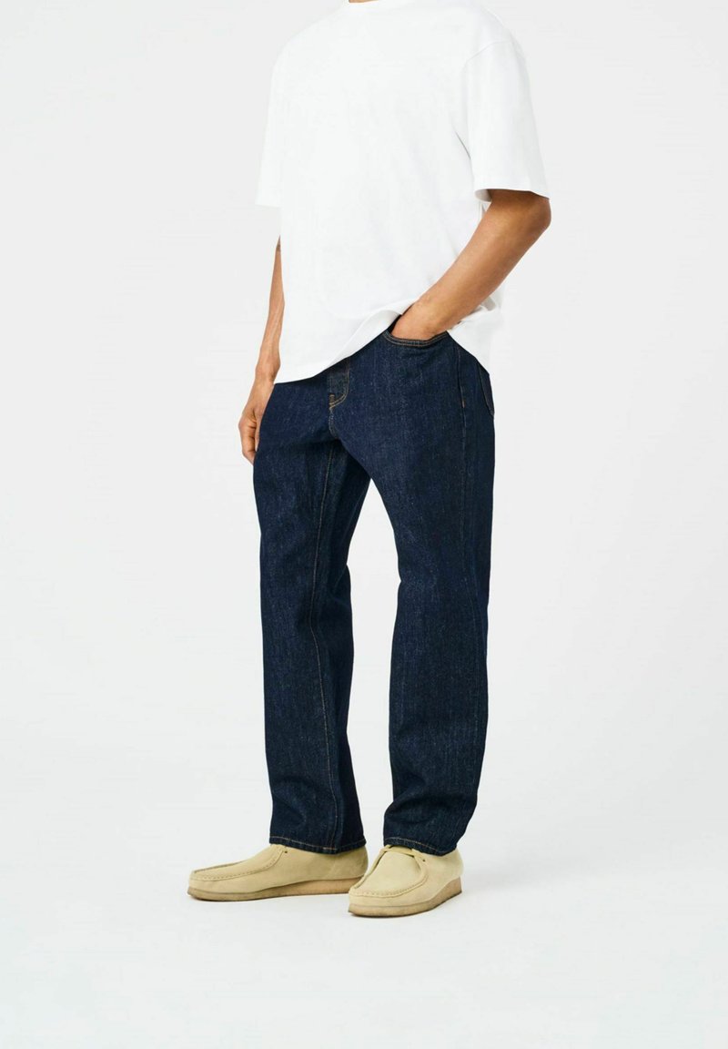 Jean en denim bleu foncé avec des jambes droites, présentant des coutures oranges et des poches latérales. Associé à un T-shirt blanc uni et des chaussures beiges.
