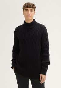 Črna pletena turtleneck pulover s kabelskim vzorcem na zgornji polovici in rebrasto teksturo na spodnji polovici, dolgimi rokavi in sproščenim krojem.