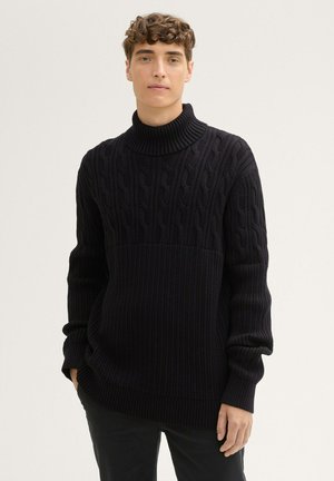 Suéter negro de punto con cuello alto que presenta un patrón de trenzas en la mitad superior y una textura acanalada en la mitad inferior, mangas largas y corte relajado.
