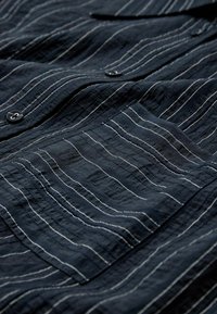 Marinblå knapop skjorte lavet af tekstureret stof med hvide vandrette striber. Har en lomme og sorte knapper. Rynket udseende.