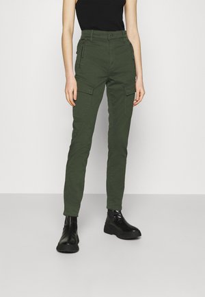 Femme portant un pantalon cargo vert foncé avec fermeture éclair et poches à rabat, un top noir sans manches et des bottines plateformes noires, debout devant un fond uni.