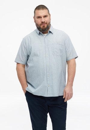 Homme avec barbe et cheveux attachés en arrière, portant une chemise bleu clair à manches courtes boutonnée et un pantalon foncé, debout avec une main dans la poche.