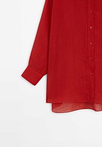 Chemise rouge à manches longues avec boutons, réalisée en tissu léger et texturé, équipée d'un ourlet arrondi et d'une poche poitrine unique.