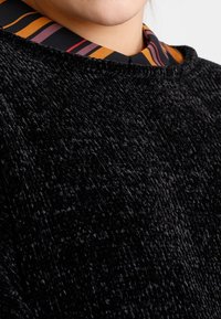 Maglione nero dalla texture morbida e pelosa con scollatura rotonda, indossato sopra un colletto di camicia a righe multicolori.