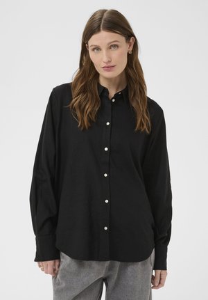 Junge Frau mit langen braunen Haaren trägt ein schwarzes Hemd mit Knöpfen und graue Hosen, steht vor einem schlichten hellgrauen Hintergrund.