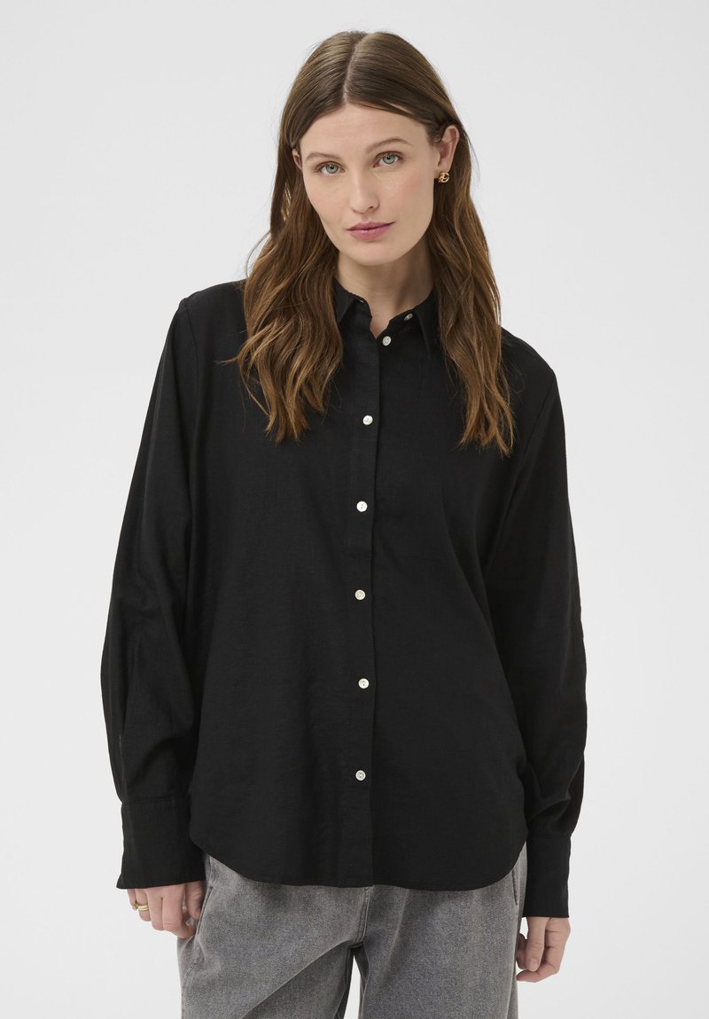 Jeune femme aux longs cheveux bruns portant une chemise noire boutonnée et un pantalon gris, debout devant un fond uni gris clair.