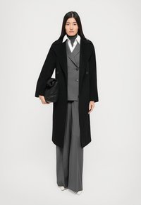Long black coat over a gray three-piece suit, featuring wide-leg pants, a white shirt, and a black textured bag. Smooth, tailored design.