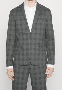 Blazer grigio a scacchi con due tasche frontali e chiusura a un bottone. Indossato sopra una camicia bianca con colletto. Tessuto dalla texture liscia.