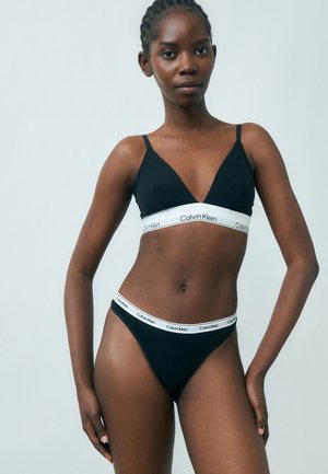 Femeie purtând un set de sutien și chiloți negri Calvin Klein cu benzi elastice albe inscripționate, stând într-un cadru simplu, cu fundal deschis la culoare.