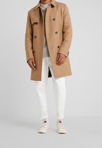 Beige Doppelt-knopf-Trenchcoat mit schwarzen Knöpfen, getragen über einem hellgrauen Pullover und weißen Hosen, kombiniert mit Sneakers im Low-Top-Stil.