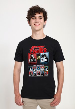 Young man smiling, wearing a black Star Wars Fighter t-shirt featuring character selection and versus graphics in red, blue, and white.