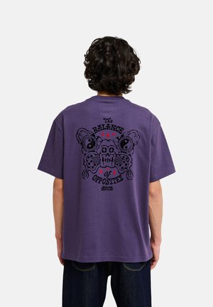 Person mit lockigem Haar trägt ein lila T-Shirt mit einem großen schwarzen Schädel- und Schmetterlingsmotiv und dem Text "The Balance of Opposites" auf dem Rücken.