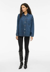 Donna con capelli ricci che indossa una giacca di denim blu oversize, pantaloni skinny neri e stivali alti neri con tacco, in piedi davanti a uno sfondo bianco.