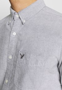 Šedá košile na knoflíky s límečkem, přední kapsou a vyšitým logem. Obsahuje bílé knoflíky a texturovaný povrch tkaniny.