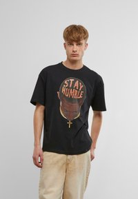 Fekete pamut póló, amelyen egy fej grafikai nyomat látható "STAY HUMBLE" felirattal és arany lánc díszítéssel. Kényelmes fazon, rövid ujjú.