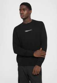 Svart hettegenser laget av mykt stoff med rund hals, lange ermer, og en rektangulær logo-patch sydd på forsiden.