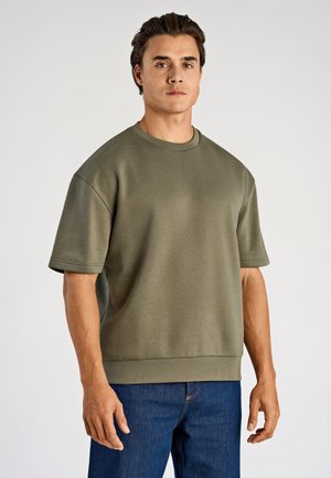 Uomo che indossa una felpa a maniche corte oversize verde oliva e jeans blu, in piedi davanti a uno sfondo chiaro e uniforme.