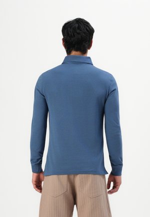 Homme de dos portant un polo bleu à manches longues et un pantalon beige sur un fond blanc uni.