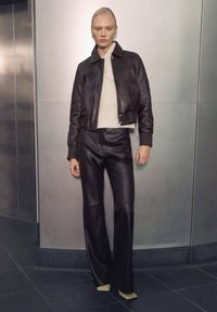 Leren cropped jacket in donkerbruin, gedragen over een lichtcrème textuurtrui, gecombineerd met wijdvallende leren broek in donkerbruin.