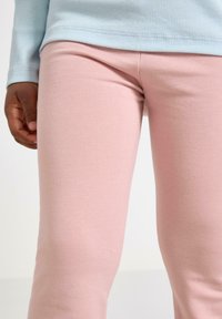 Lyserøde leggings lavet af blødt, strækbart stof, tætsiddende i taljen med en glat tekstur. En lyseblå langærmet skjorte er delvist synlig.