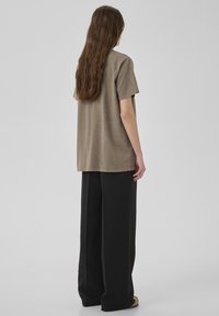 Person mit langen braunen Haaren steht mit dem Rücken zur Kamera, trägt ein lockeres braunes T-Shirt, weite schwarze Hosen und beigen Schuhe vor einem grauen Hintergrund.