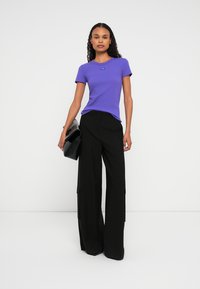 Donna che indossa un top fitted viola a maniche corte, pantaloni neri a gamba larga e tiene una borsa a tracolla nera su uno sfondo bianco uniforme.