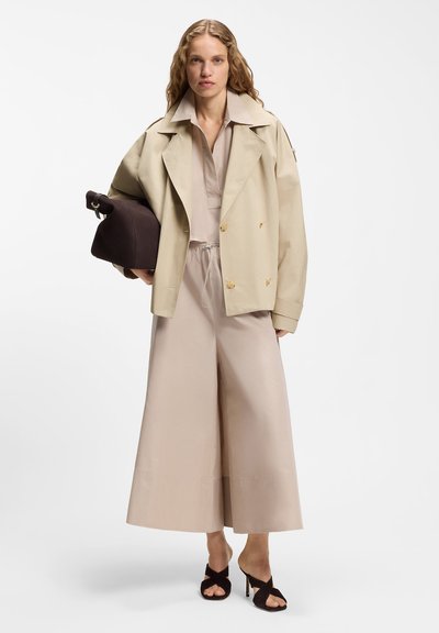 Femme portant un pantalon beige à jambes larges, une chemise assortie, une veste beige clair, des sandales à talons hauts noires, tenant un grand sac à main marron foncé.