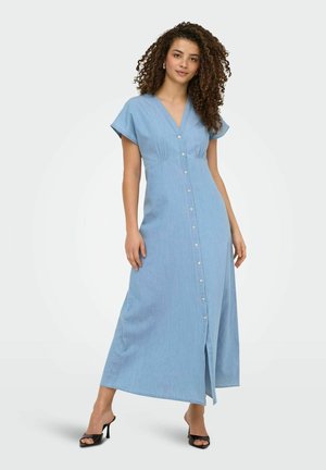 Vrouw met krullend haar, gekleed in een lichtblauwe maxi-jurk met knopen en zwarte sandalen met hak, staand tegen een witte achtergrond.