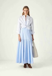 Ανοικτό μπλε μακρύ φόρεμα με ραμμένο φούστα σε επίπεδα, σε συνδυασμό με λευκό πουκάμισο δεμένο και καφέ μπότες αστραγάλου. Περιλαμβάνει μία μπεζ τσάντα.