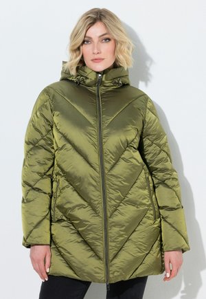 Donna con capelli biondi che indossa una giacca imbottita trapuntata verde oliva con cappuccio, in piedi davanti a uno sfondo chiaro uniforme.