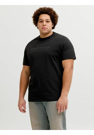 Göndör hajú férfi fekete "ORIGINALS" feliratú pólóban és világoskék farmerral, sima világos háttér előtt állva.