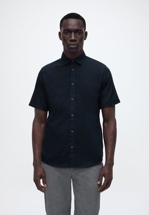 Férfi fekete rövid ujjú, gombos ingben és szürke nadrágban, fehér, egyszínű háttér előtt áll, előre nézve.