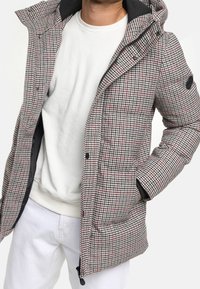 Harmaa ja punainen ruudullinen toppatakki hupulla, jossa painonapit ja sivutaskut, puettu valkoisen neuleen ja vaaleiden housujen päälle.