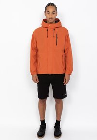 Oranje waterdichte jas met een capuchon, ritszak op de borst en zijzakken. Voorzien van elastische manchetten en een verstelbare trekkoord onderaan.