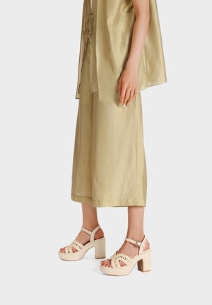 Sandali platform beige con dettagli intrecciati, punta aperta, cinghietta alla caviglia e tacco a blocco texturizzato, abbinati a un outfit leggero in oro metallico.