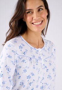 Top con estampado floral azul sobre un fondo blanco, con cuello redondo, cierre de botones y detalles de piping azul claro.