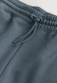 Gros plan sur la taille d’un pantalon de jogging bleu-gris avec bande élastique, cordon noué et couture visible de la poche latérale.
